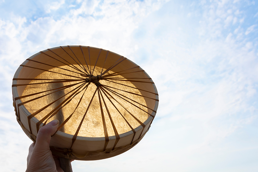 Eine Hand hält eine traditionelle Rahmentrommel gegen den hellen Himmel, durch den das Sonnenlicht die gespannte Tierhaut durchscheinen lässt.
