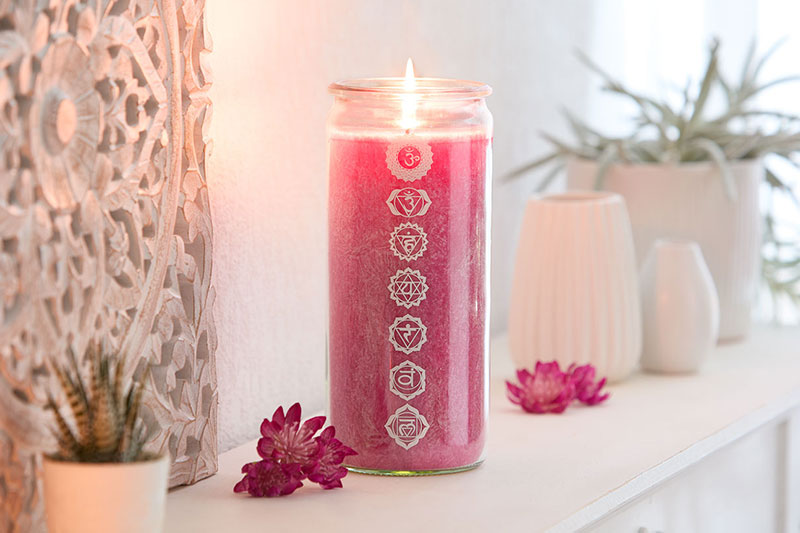 Große magentafarbene Kerze im Glas mit sieben weißen Chakra-Symbolen, brennend auf einem weißen Regal, dekoriert mit magentafarbenen Blüten.