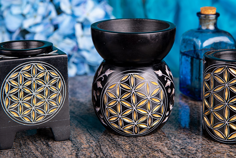 Verschiedene Aromalampen, alle schwarz mit goldener Blume des Lebens, stehen auf einer Tischplatte vor türkisblauem Hintergrund.