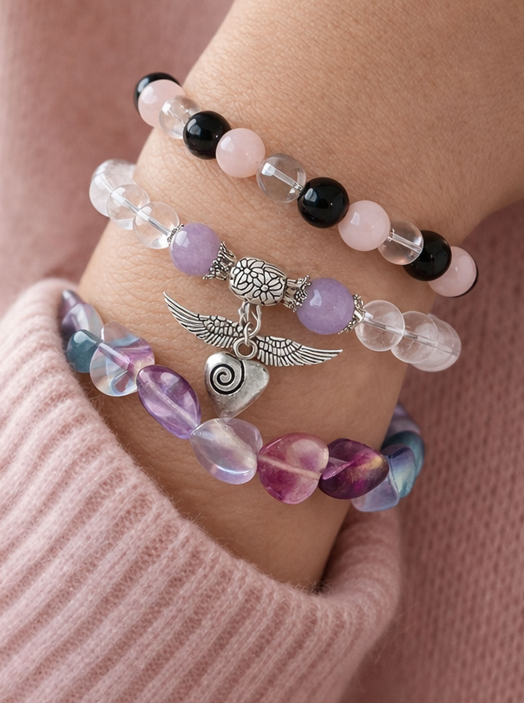 Hand in rosafarbenem Pullover trägt mehrere filigrane Armbänder mit bunten und zarten Perlen sowie Anhängern, sanft beleuchtet, wirkt harmonisch und feminin.