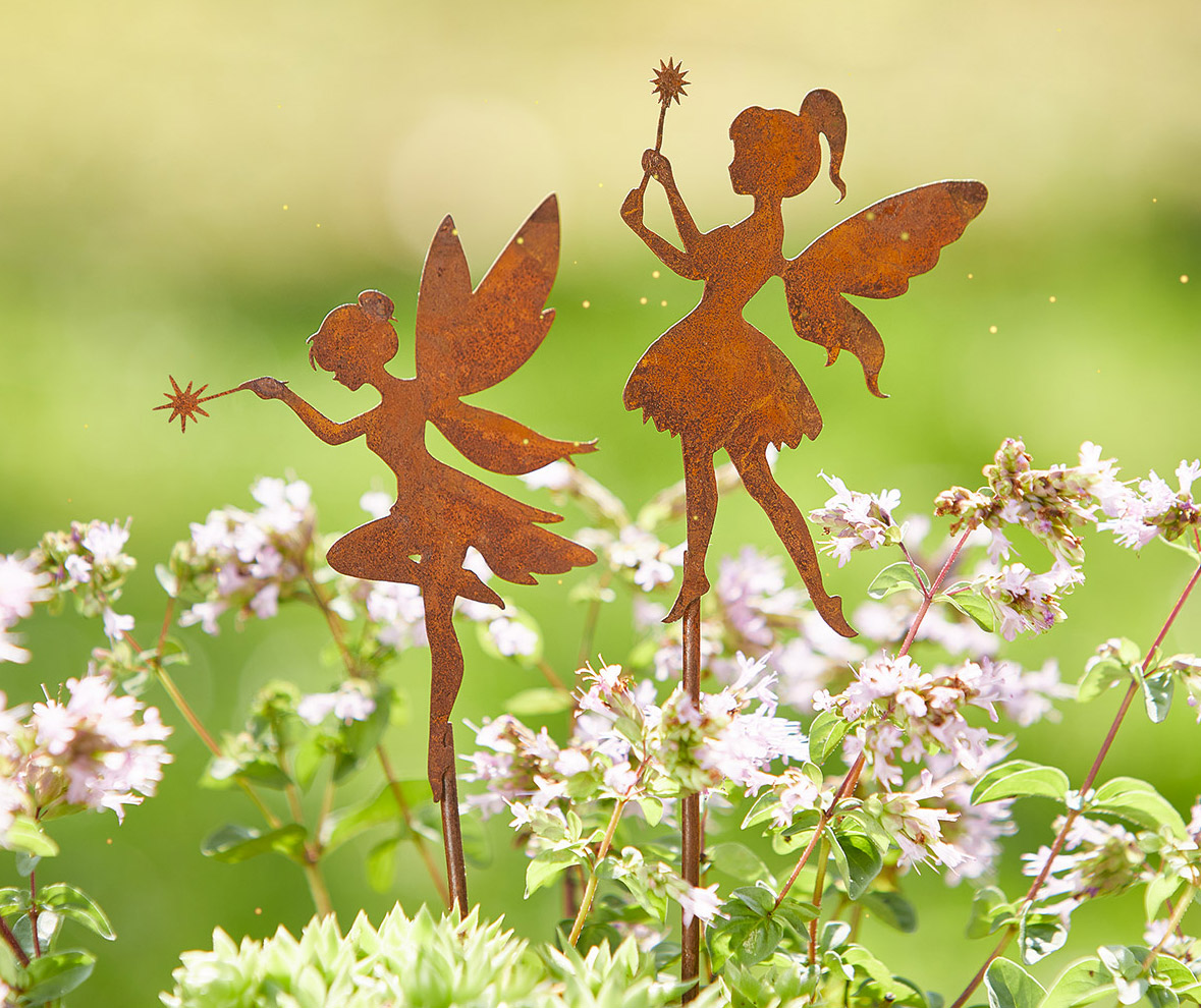 Zwei rostfarbene Elfenfiguren aus Metall mit Zauberstäben stehen zwischen blühenden Kräutern und zarten Blüten vor einem weich verschwommenen, sonnigen Gartenhintergrund.