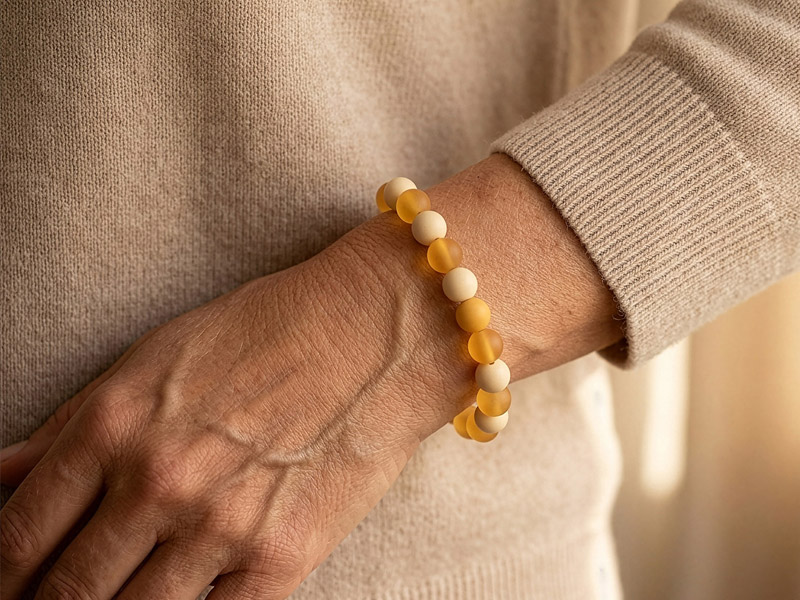 Hand mit warmem, beigefarbenem Pullover trägt ein Weihrauch-Armband aus gelben und cremefarbenen Perlen, sanftes Licht betont die natürliche, ruhige Ausstrahlung.