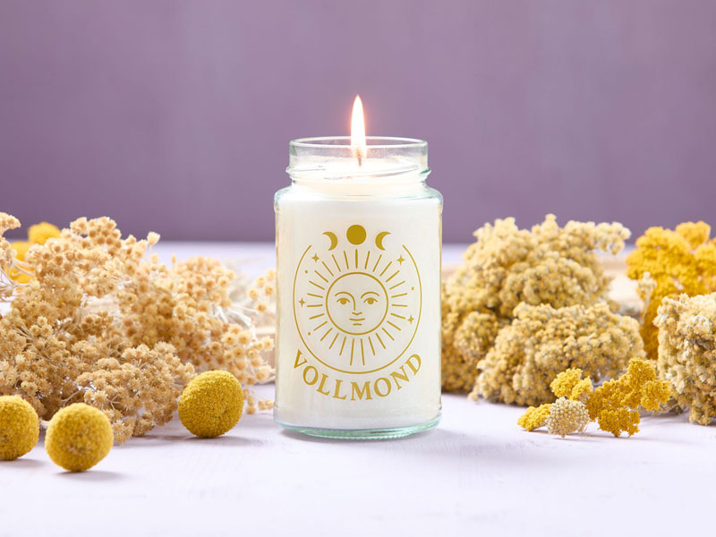 Weiße Duftkerze in einem schlichten Glas mit goldfarbener Sonnen- und Mondsymbolik sowie dem Schriftzug „Vollmond“, liebevoll arrangiert zwischen Trockenblumen.