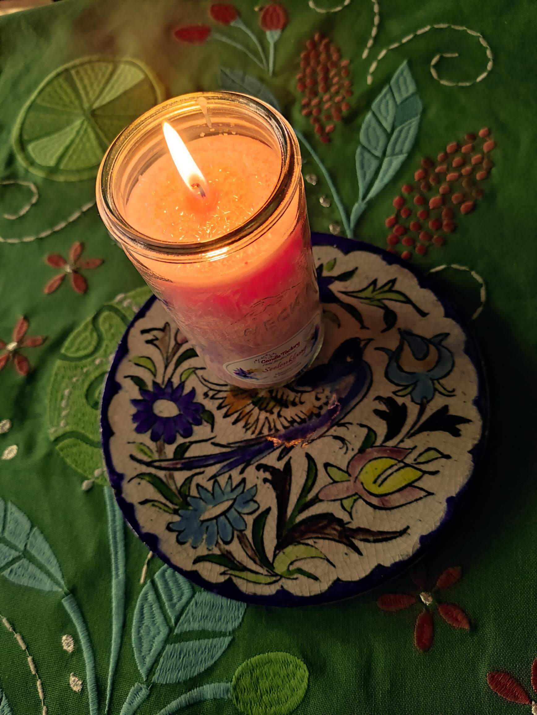 Brennende Kerze auf einem Teller mit floralem Muster, der auf einer grünen Tischdecke mit Stickereien steht