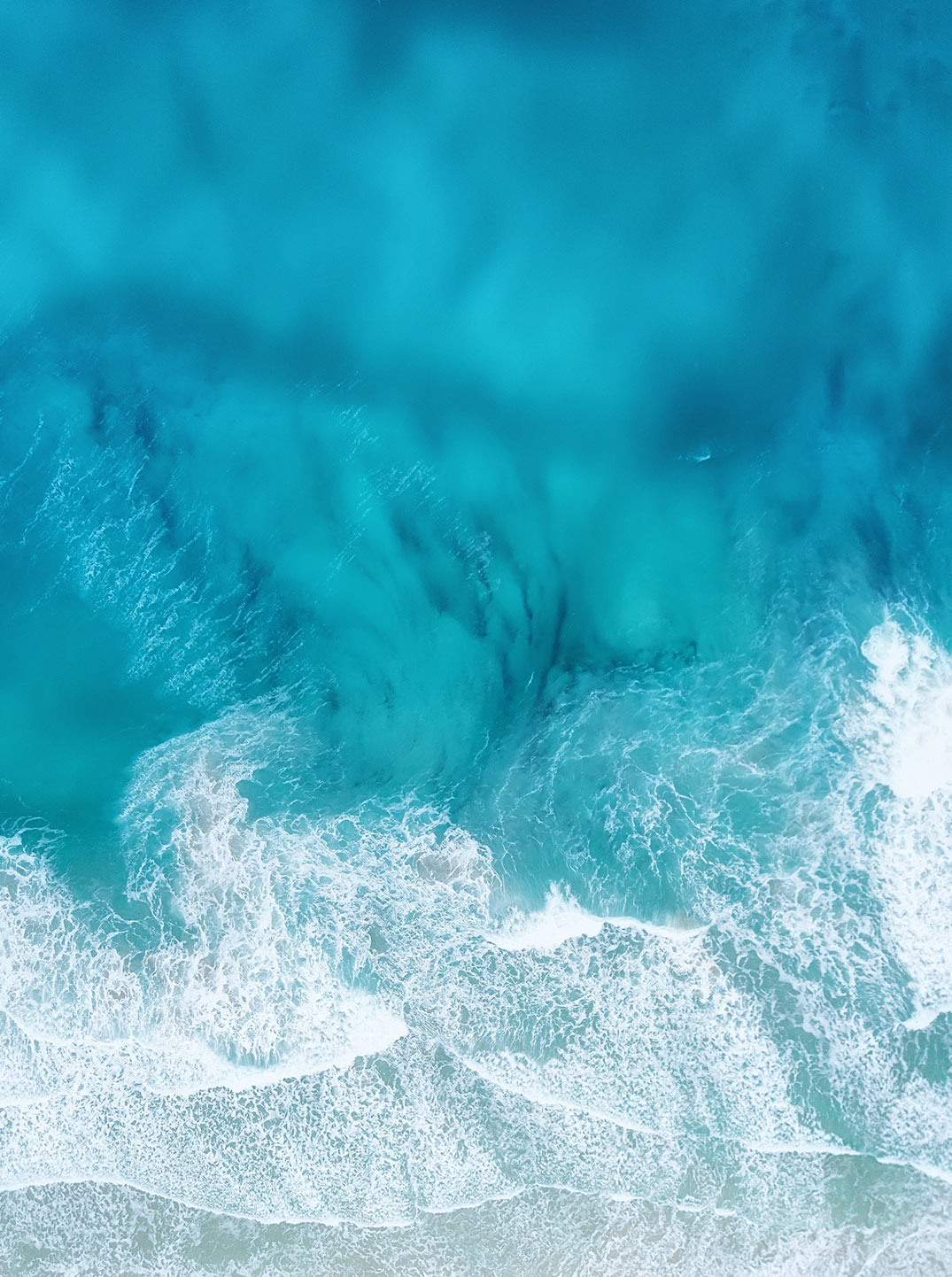 Luftaufnahme von türkisblauem Meerwasser, das in sanften Wellen an einen hellen Sandstrand brandet.