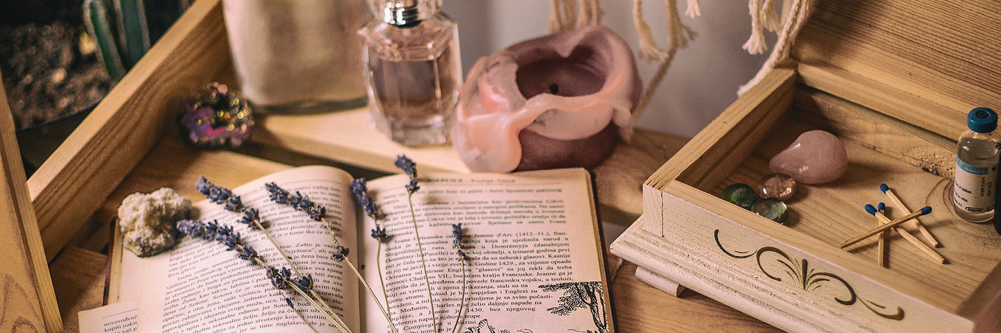 Auf einem Holztisch sind verschiedene Gegenstände angeordnet: Ein Buch, eine Kerze, ein Parfüm-Flakon, mehrere bunte Halbedelsteine, eine Holzschatulle sowie getrockneter Lavendel.