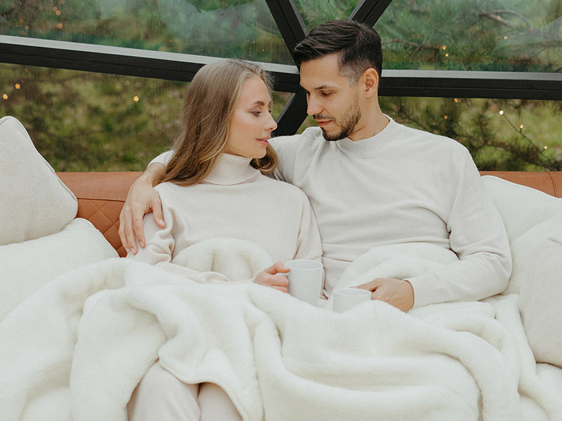 Ein Paar sitzt auf einem Sofa, eingekuschelt in eine weiße dicke Decke aus Merino-Kaschmir-Wolle