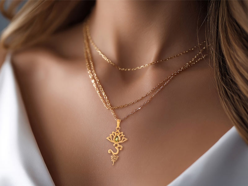 Zart glänzender goldener Anhänger mit kunstvollem Lotus- und Om-Symbol, getragen an einer filigranen Doppel-Halskette. Das Schmuckstück liegt sanft auf der Haut einer Frau in einem weißen, tief ausgeschnittenen Oberteil.