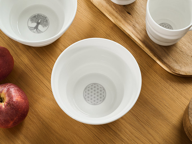 Mehrere weiße edle Porzellanschalen und -becher stehen auf einem hellen Holztisch. In den Böden der Gefäße sind in leuchtendem Silber der Lebensbaum oder die Blume des Lebens eingearbeitet.