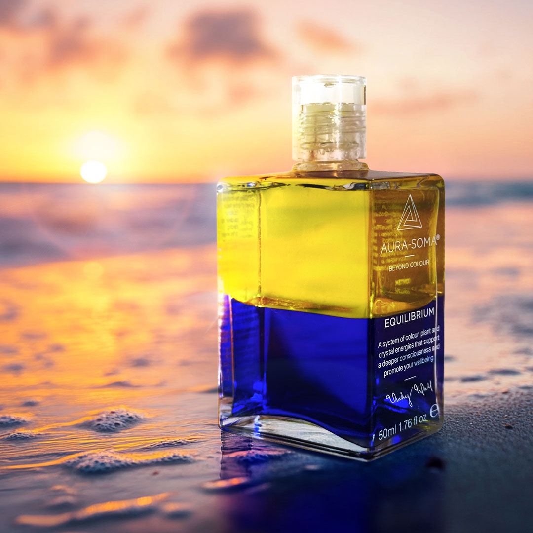 Aura Soma Equilibrium B123: Zitronengelb über Königsblau; Die Flasche steht auf einem Sandstrand, im Hintergrund ist ein leuchtender Sonnenaufgang zu sehen.