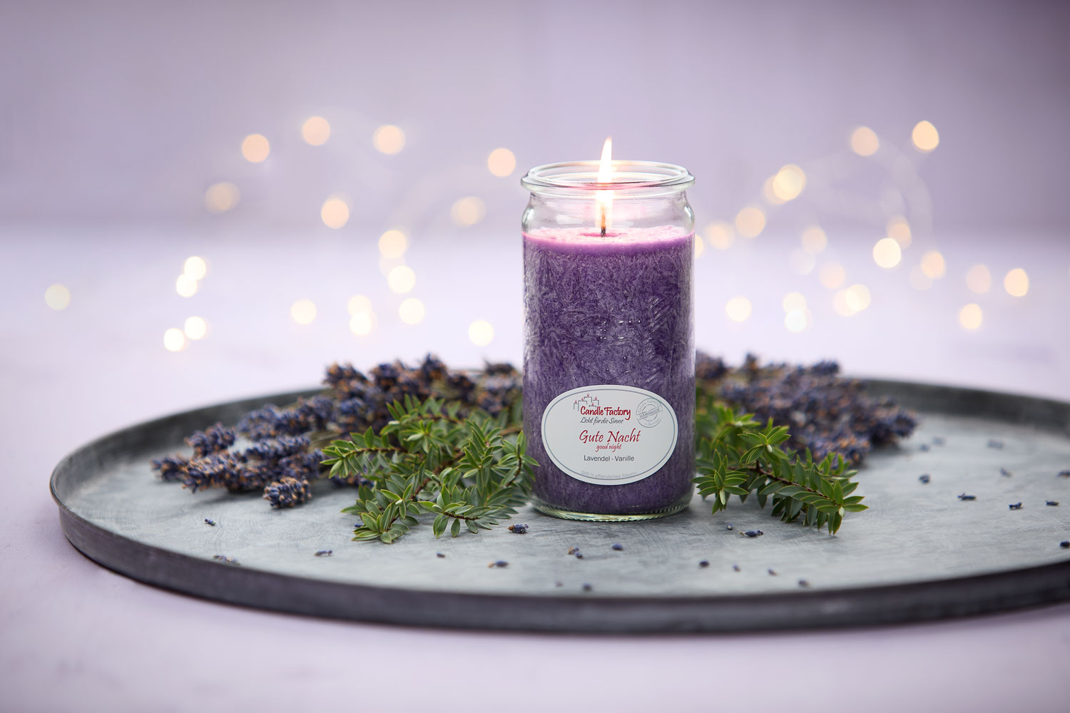 Lila Duftkerze mit Etikett „Gute Nacht“, umgeben von Lavendelblüten und Grün auf einem Metalltablett. Im Hintergrund zarte Lichtpunkte – stimmungsvolle Szene für Abendruhe und Entspannung.