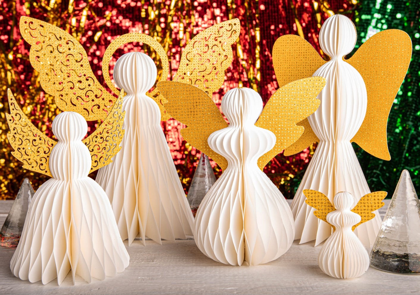 Weiße und goldene Papierengel in verschiedenen Größen stehen vor einem festlich glitzernden Hintergrund in Rot, Gold und Grün.
