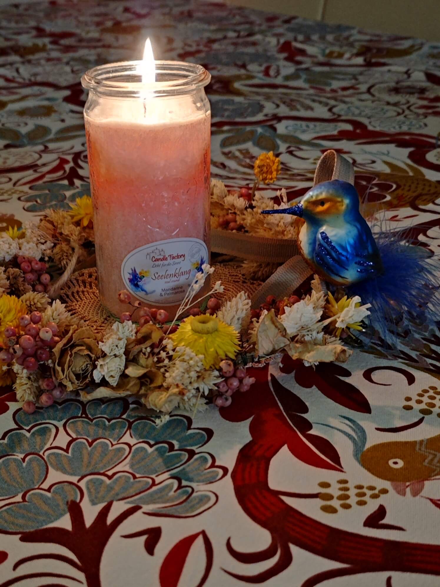 Brennende Kerze in einem Blumekranz mit Eisvogel-Figur auf einer bunten Tischdecke mit floralem Muster