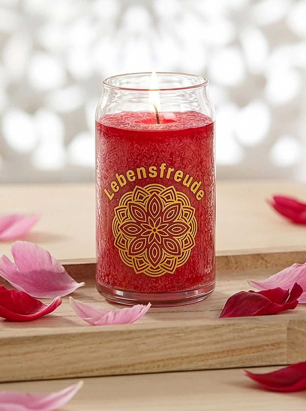 Rote Kerze im Glas mit der Aufschrift „Lebensfreude“, umgeben von zarten rosa Blütenblättern auf einem Holztablett, stimmungsvoll in weichem Licht arrangiert.