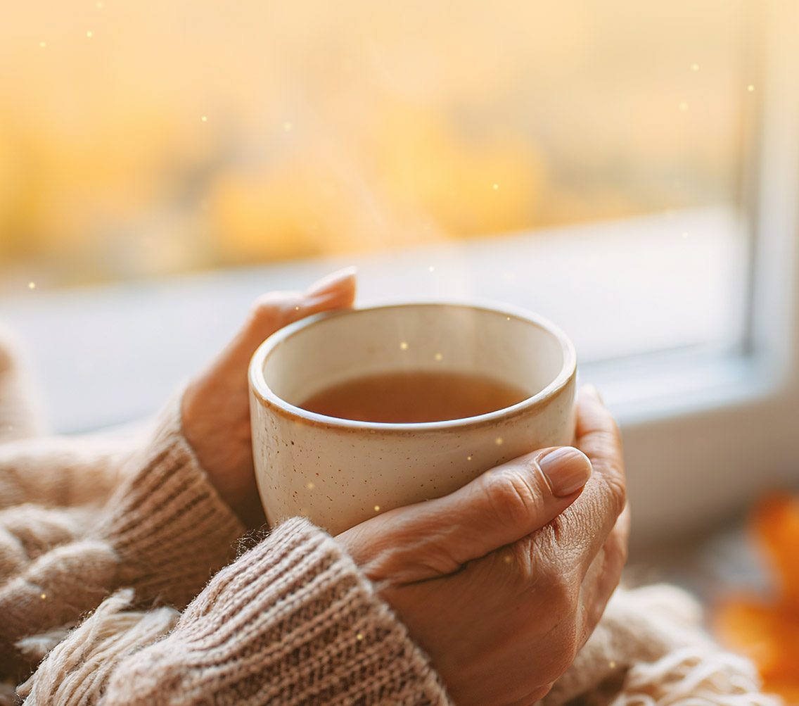 In ein weiches Plaid gehüllte Hände halten eine dampfende Tasse Tee, im Hintergrund goldene Herbstblätter. Die Szene ist in warmes Licht getaucht.