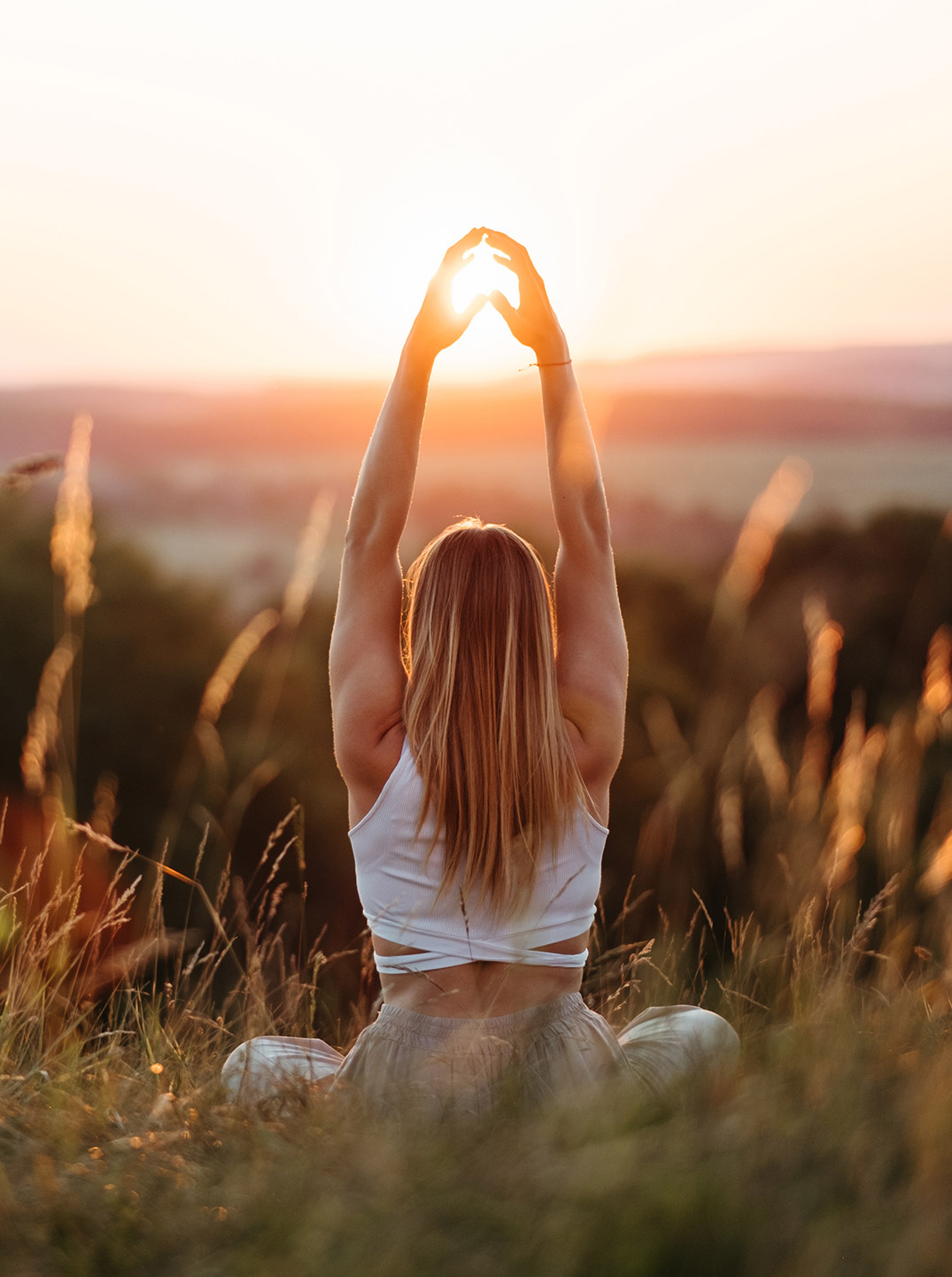 Frau sitzt im hohen Gras auf einem Hügel bei Sonnenuntergang und streckt die Arme zur Sonne empor, mit weiter Landschaft im warmen Abendlicht im Hintergrund.