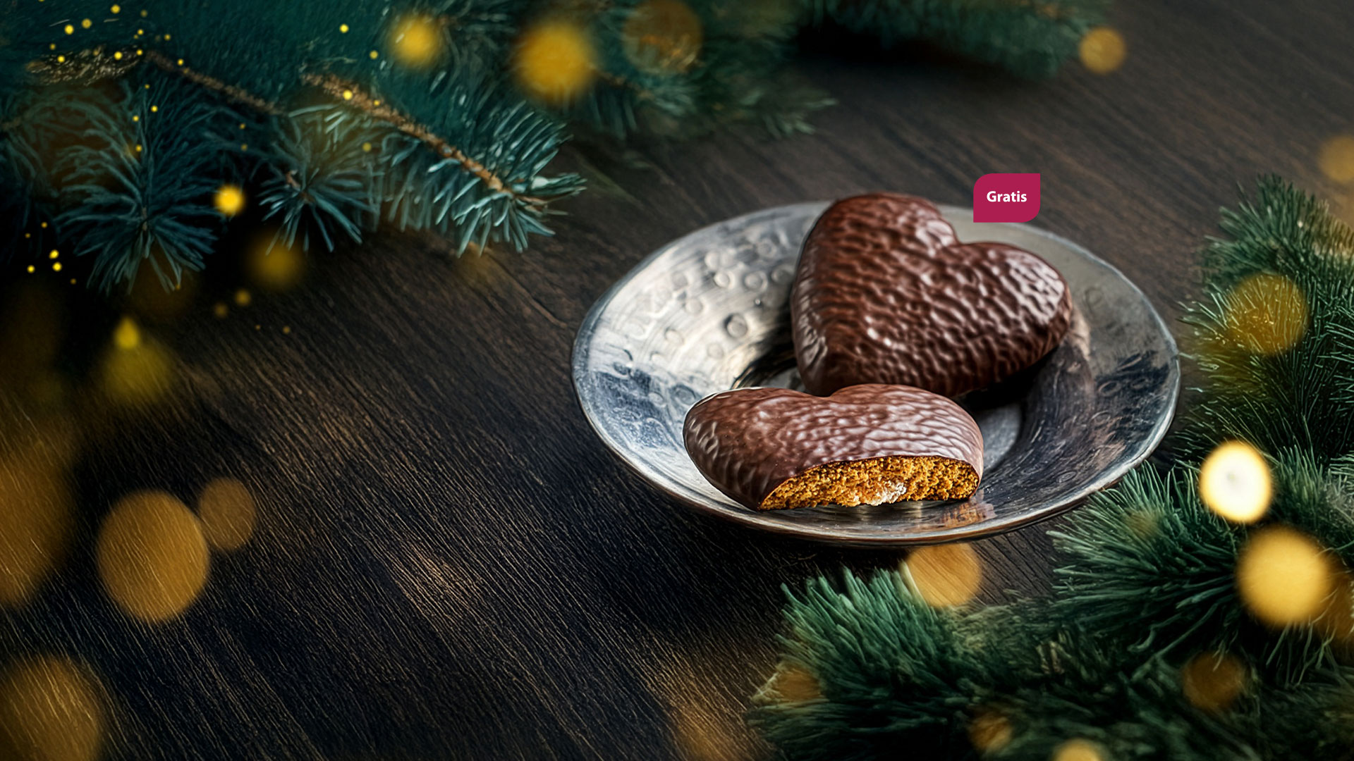 Festliche Szene mit zwei schokolierten Lebkuchen-Herzen in einer silbernen Schale, umrahmt von Tannenzweigen und goldenem Lichterglanz.