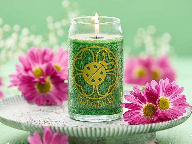 Grüne Duftkerze im Glas mit goldfarbener Glückssymbolik (Marienkäfer im Kleeblatt), brennend zwischen pinken Blüten auf einem hellen Teller – ein stimmungsvolles Arrangement für gute Wünsche und Neuanfang.