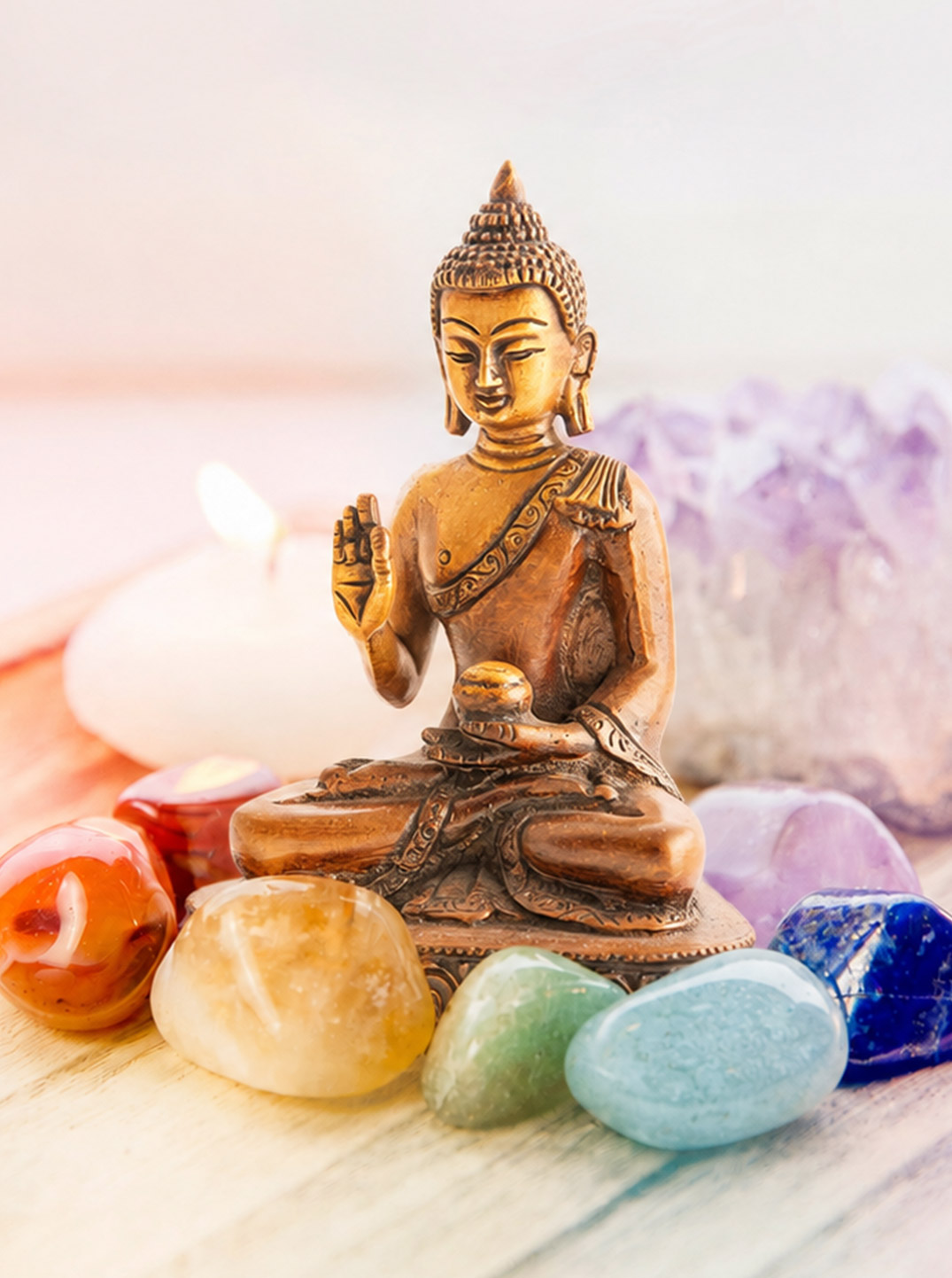Kleine Buddha-Figur sitzt auf einem hellen Holztablett, umgeben von bunten Trommelsteinen und einem Amethyst-Cluster. Mehrere weiße Teelichter sorgen für eine ruhige, meditative Atmosphäre vor hellem Hintergrund.