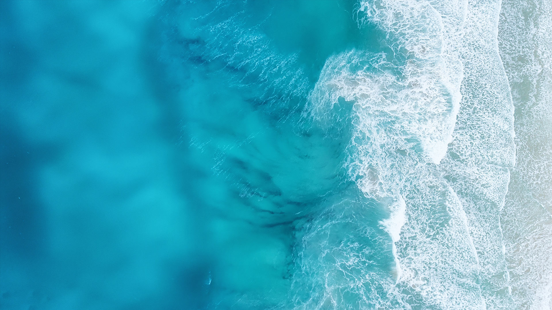 Luftaufnahme von türkisblauem Meerwasser, das in sanften Wellen an einen hellen Sandstrand brandet.