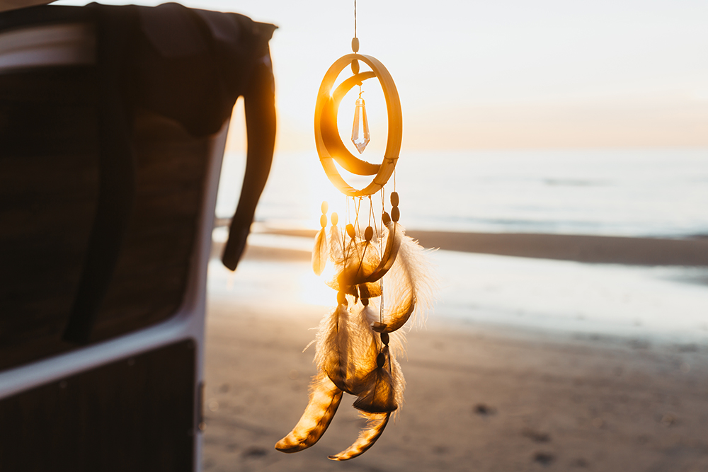 Ein Traumfänger mit Federn und Kristallanhänger hängt an einem Fahrzeug und wird von der warmen Abendsonne am Strand beleuchtet.