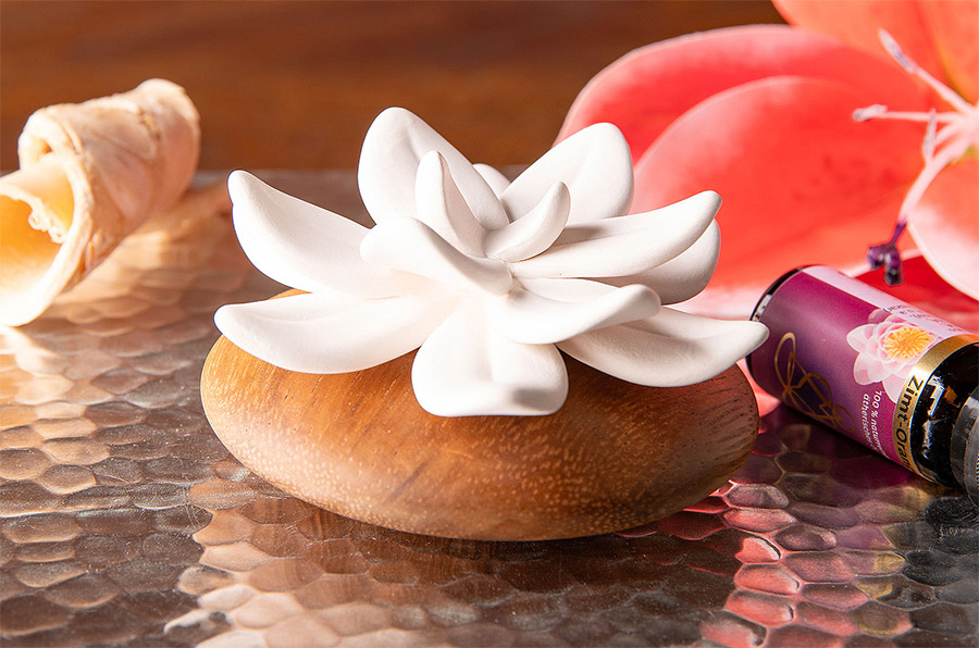 Ein zart geformter Duftstein in Form einer weißen Blüte ruht auf einem runden Holzsockel. Daneben liegen ein violettes Fläschchen mit ätherischem Öl.