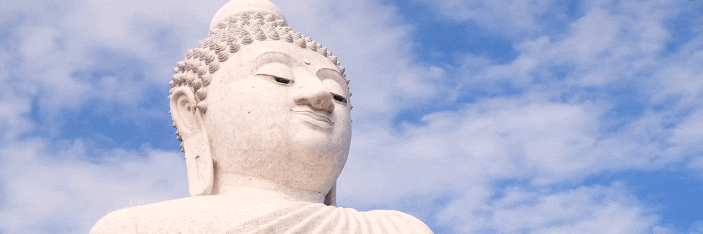 Gesicht einer großen, steinernen Buddha-Statue, im Hintergrund ziehen weiße Wolken über den blauen Himmel
