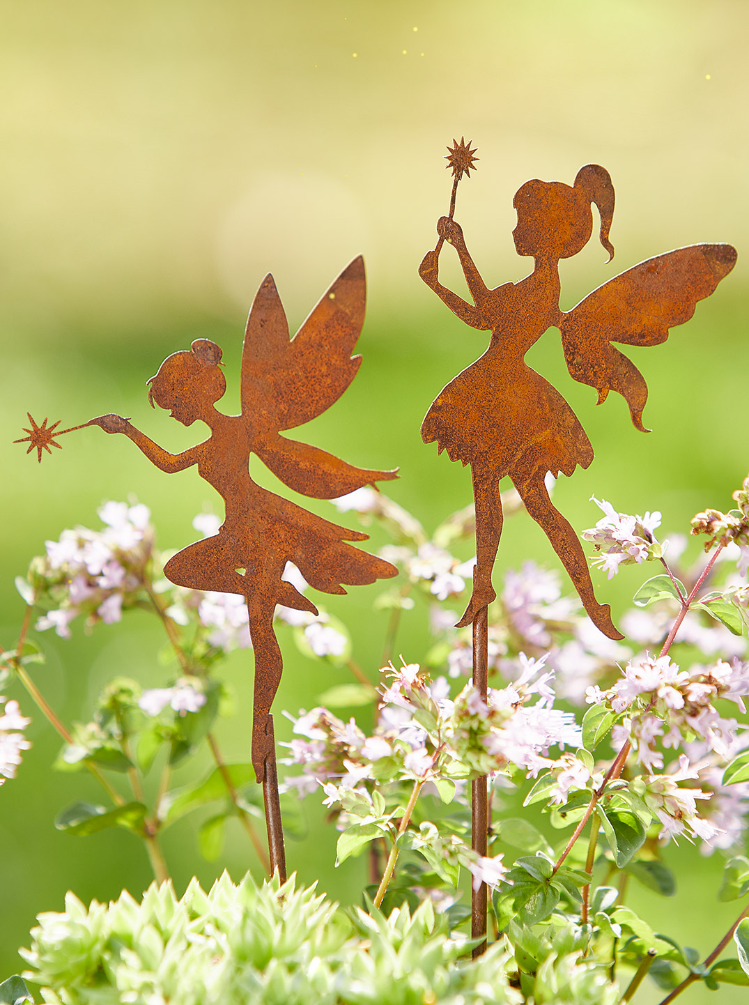 Zwei rostfarbene Elfenfiguren aus Metall mit Zauberstäben stehen zwischen blühenden Kräutern und zarten Blüten vor einem weich verschwommenen, sonnigen Gartenhintergrund.