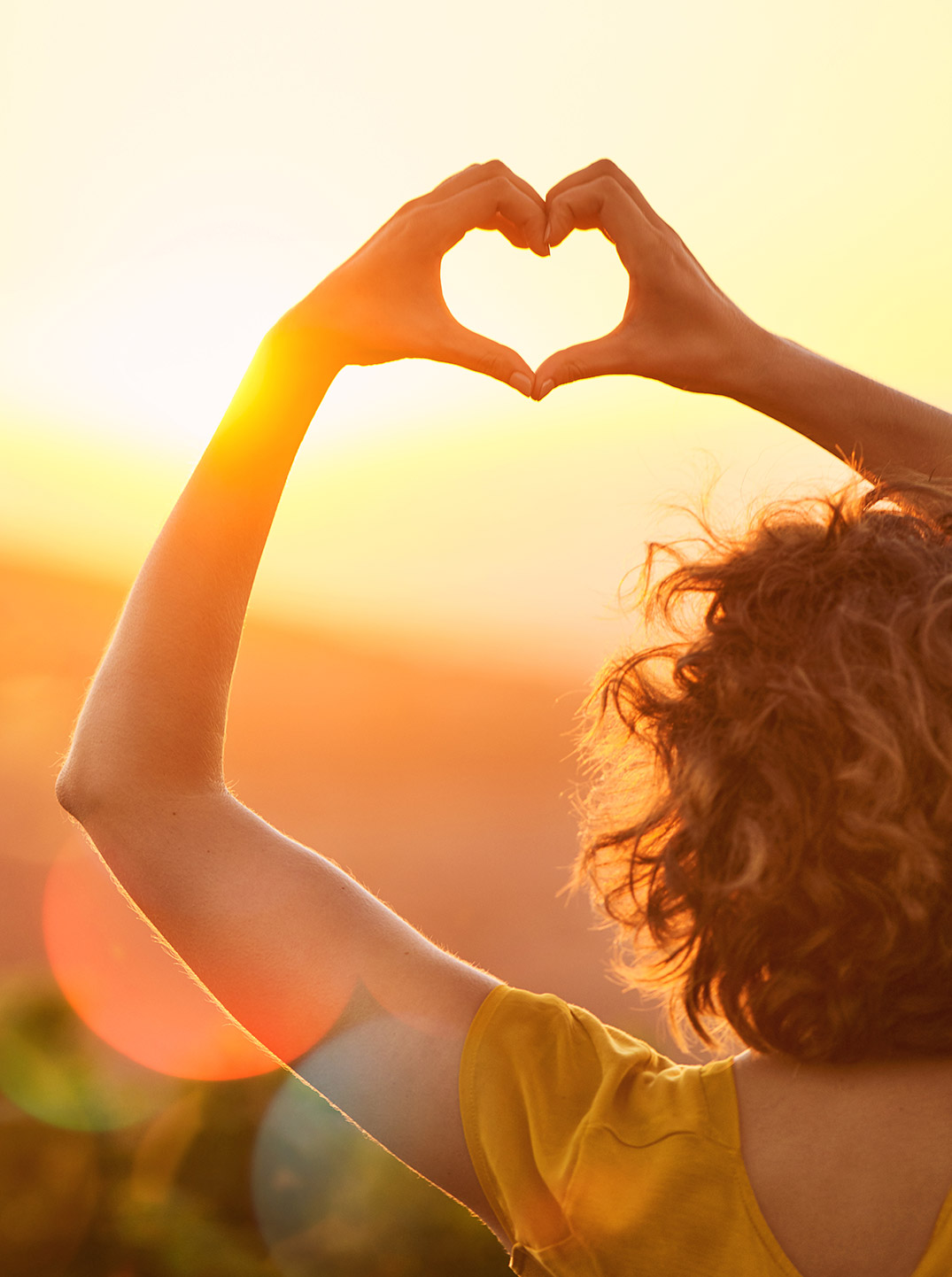 Person blickt in den Sonnenuntergang und formt mit den Händen ein Herz vor der tief stehenden Sonne. Warmes goldenes Licht, sanfte Hügel und weiche Lichtreflexe vermitteln ein Gefühl von Lebensfreude, Dankbarkeit und Verbundenheit.