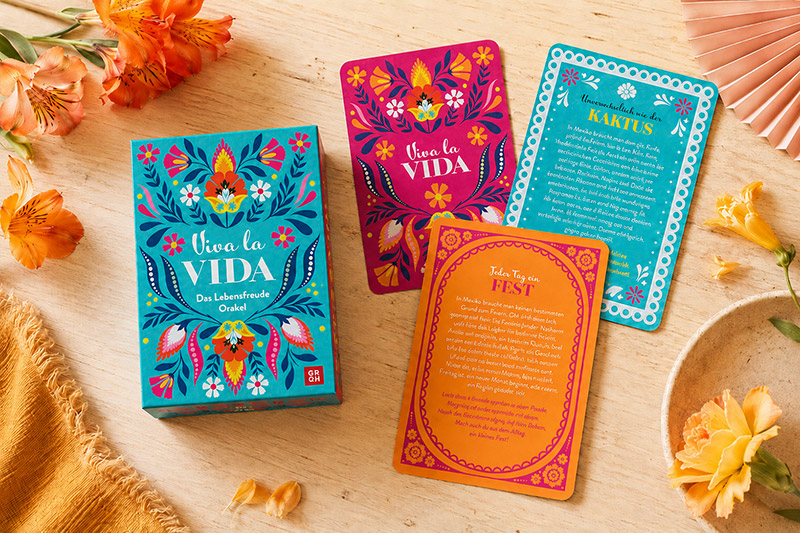 „Viva la Vida – Das Lebensfreude Orakel“ liegt mit drei ausgelegten Karten auf einem hellen Holztisch. Die Karten mit Blumenmuster in Türkis, Pink und Orange sowie frische Blüten schaffen eine fröhliche, mexikanisch inspirierte Atmosphäre.