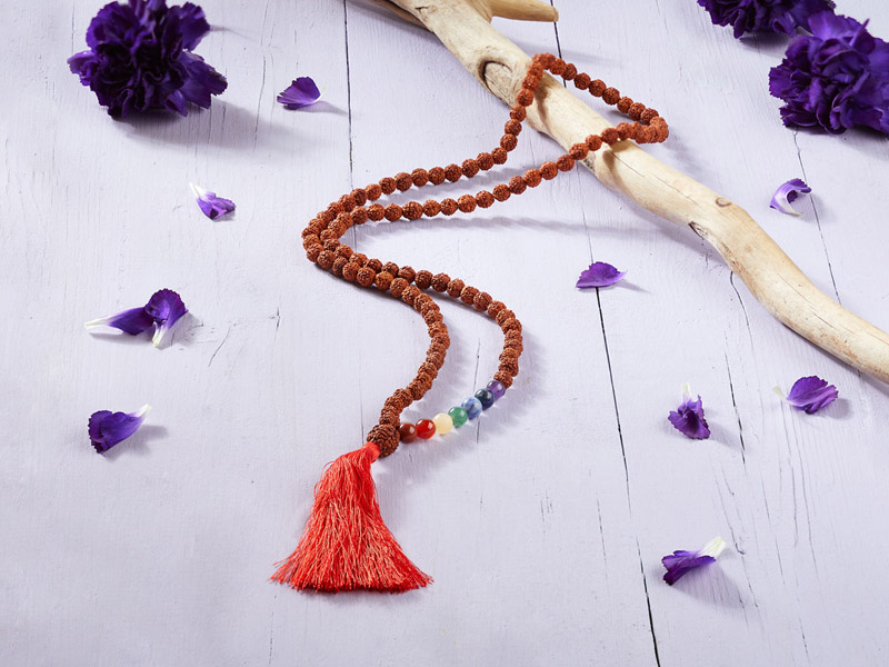 Rudraksha-Mala mit roten Quasten und bunten Chakra-Perlen liegt auf einer hellen Holzfläche, umgeben von violetten Blütenblättern und einem dekorativen Ast.