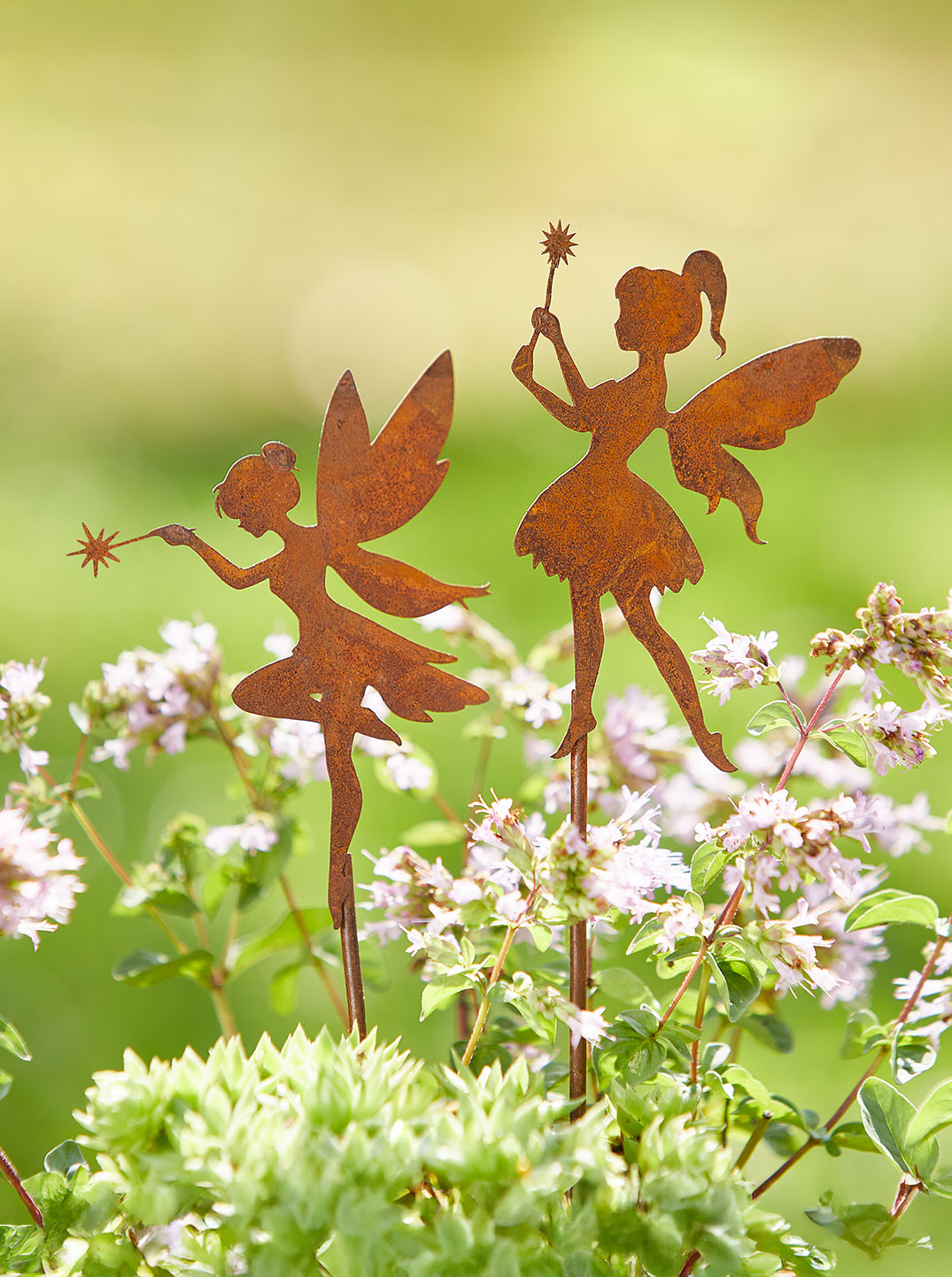 Zwei Edelrost-Gartenstecker in Form von Feen stehen zwischen blühenden Kräutern. Beide Feen haben feine Flügel und halten einen Zauberstab mit sternförmiger Spitze. In einem Kreis daneben steht "-15%".