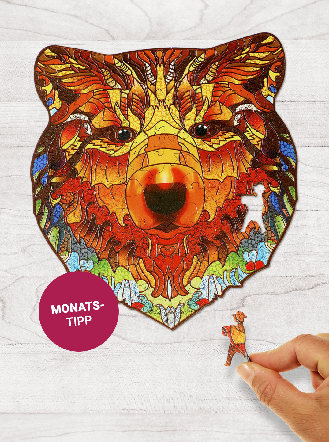 Ein buntes Holzpuzzle in Form eines Bärenkopfes liegt auf einem hellen Holzuntergrund. Daneben steht "Monatstipp".