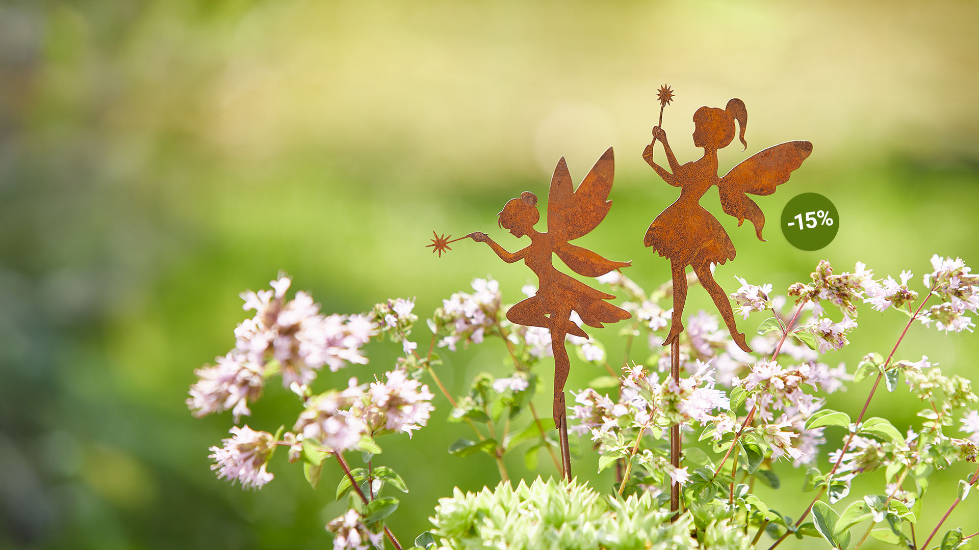 Zwei Edelrost-Gartenstecker in Form von Feen stehen zwischen blühenden Kräutern. Beide Feen haben feine Flügel und halten einen Zauberstab mit sternförmiger Spitze. In einem Kreis daneben steht "-15%".