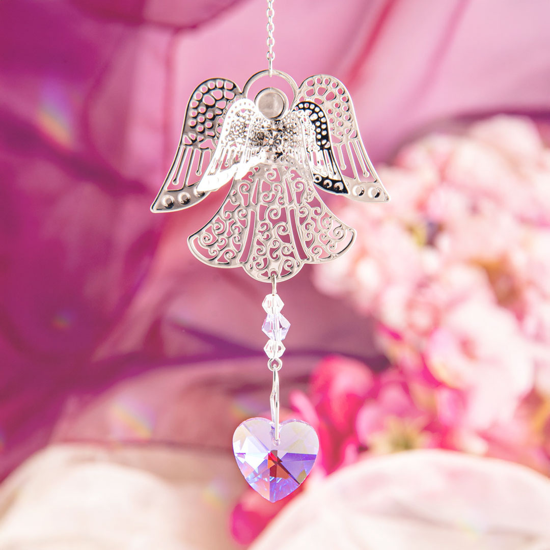 Ein filigraner Sonnenfänger in Form eines Engels hängt vor einem rosa Hintergrund mit Blumen. An seinem unteren Ende sind transparente Kristallperlen und ein schimmerndes Herz aus facettiertem Glas angebracht, das in zarten Regenbogenfarben funkelt. 