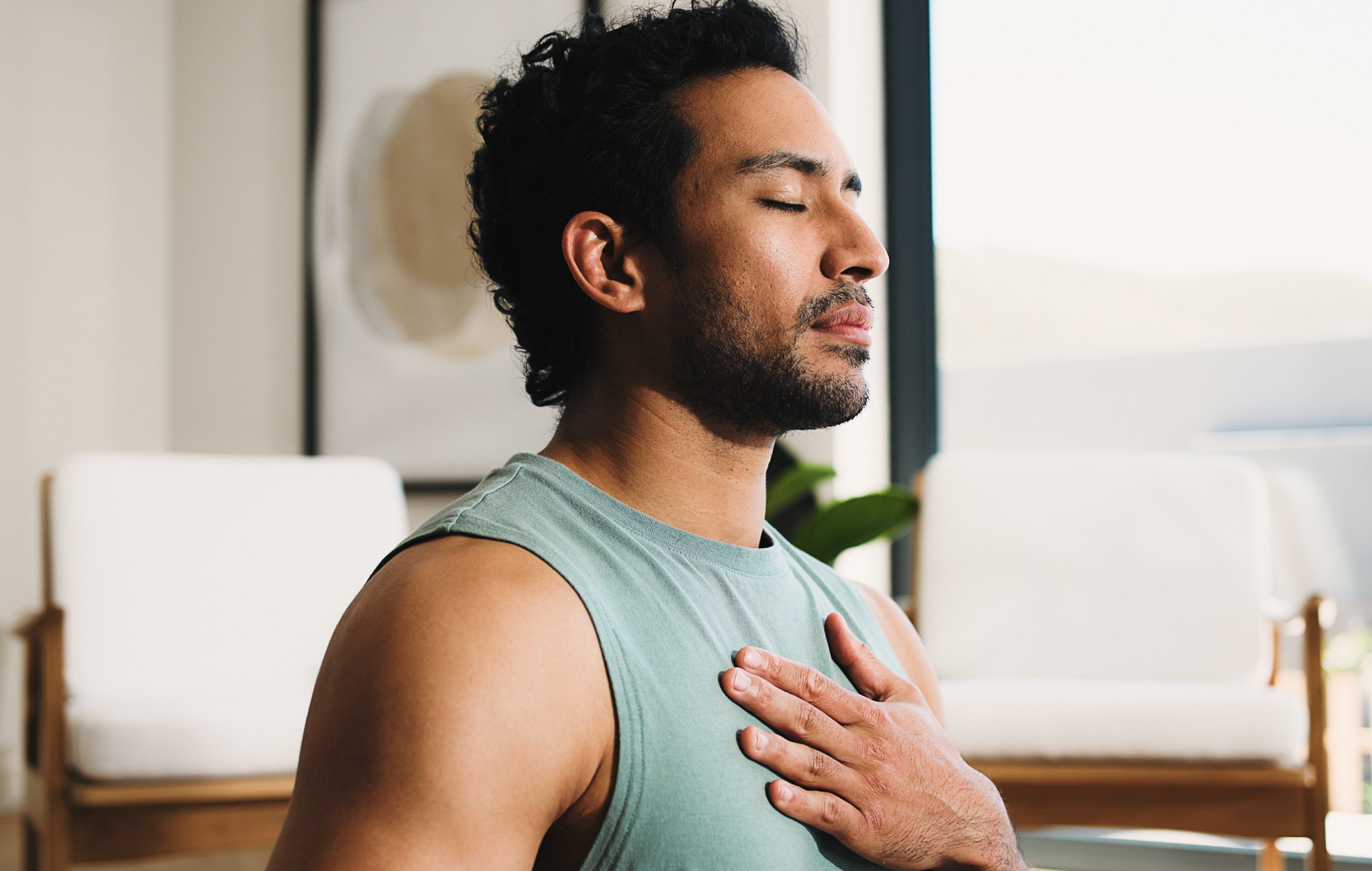 Mann mit geschlossenen Augen legt die Hand auf die Brust und atmet entspannt in einem hellen Raum.