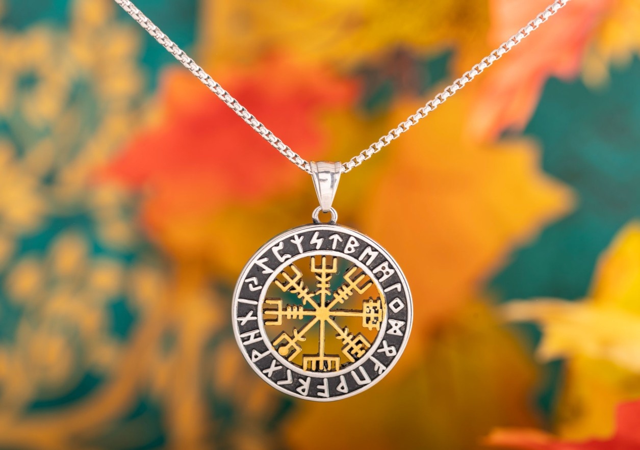 Silberfarbene Halskette mit rundem Anhänger, der ein goldfarbenes nordisches Vegvísir-Symbol und Runenzeichen zeigt, vor einem farbenfrohen, unscharfen Hintergrund.