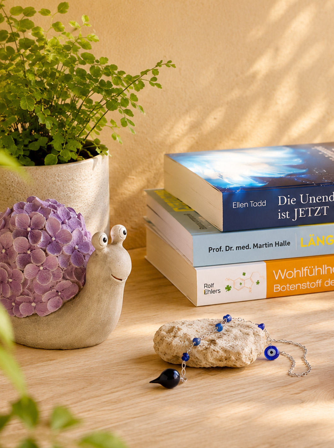 Auf einem hellen Holztisch liegt ein Stapel mit drei Büchern. Daneben stehen eine brennende rosa Duftkerze und ein Räucherstäbchenhalter. Eine Schneckenfigur und ein Pendel ergänzen die sommerliche Szene.