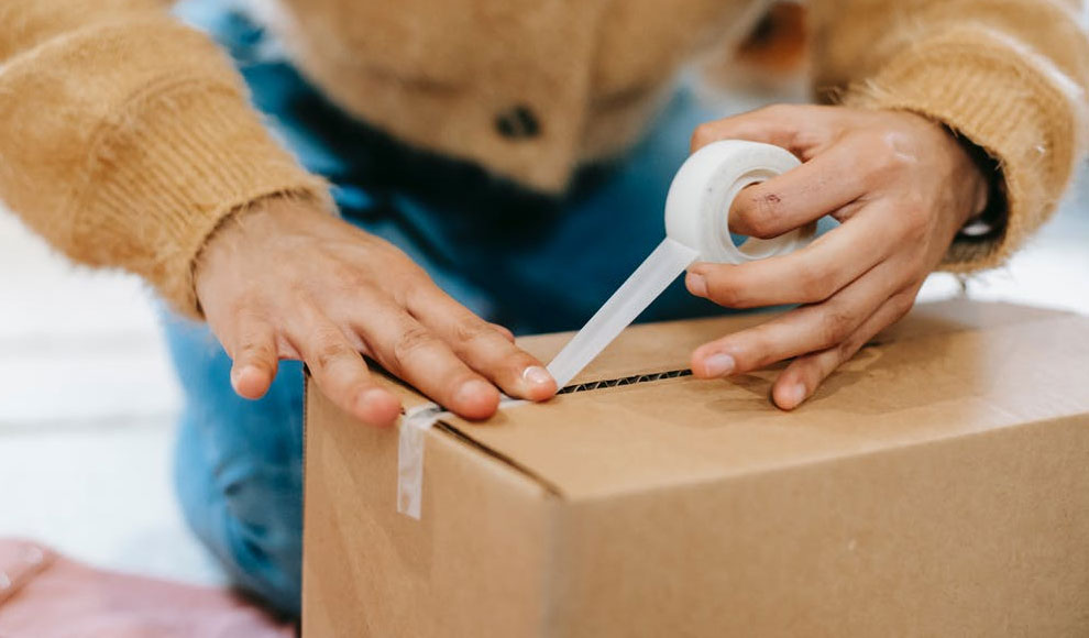 Eine Person kniet hinter einem Paket und verschließt es mit Klebeband.