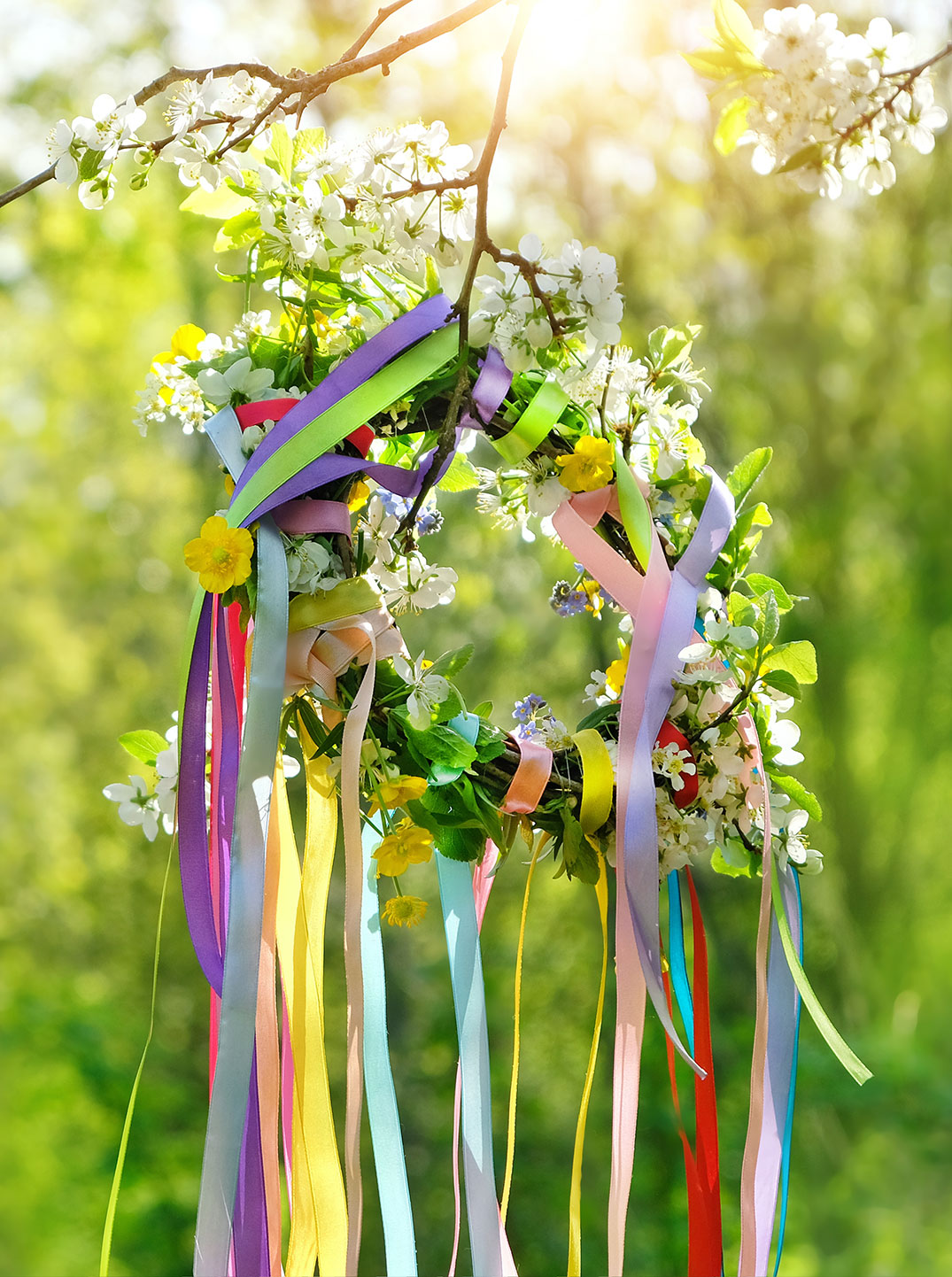 Bunter Frühlingskranz mit Blumen und langen, farbigen Bändern hängt an einem blühenden Zweig im Sonnenlicht vor einem grünen, weichen Hintergrund.