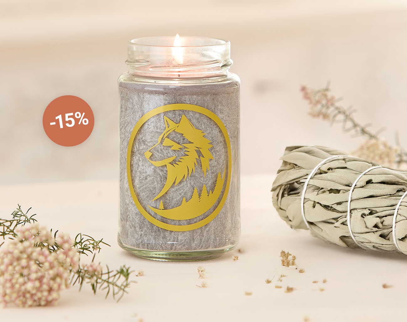 Eine graue Kerze in einem Glasgefäß mit goldfarbenem Wolfssymbol brennt auf einem hellen Tisch. Daneben liegt ein Bündel weißer Salbei und zarte Blütenzweige. Ein runder Hinweis mit '-15%' zeigt eine Rabattaktion an.