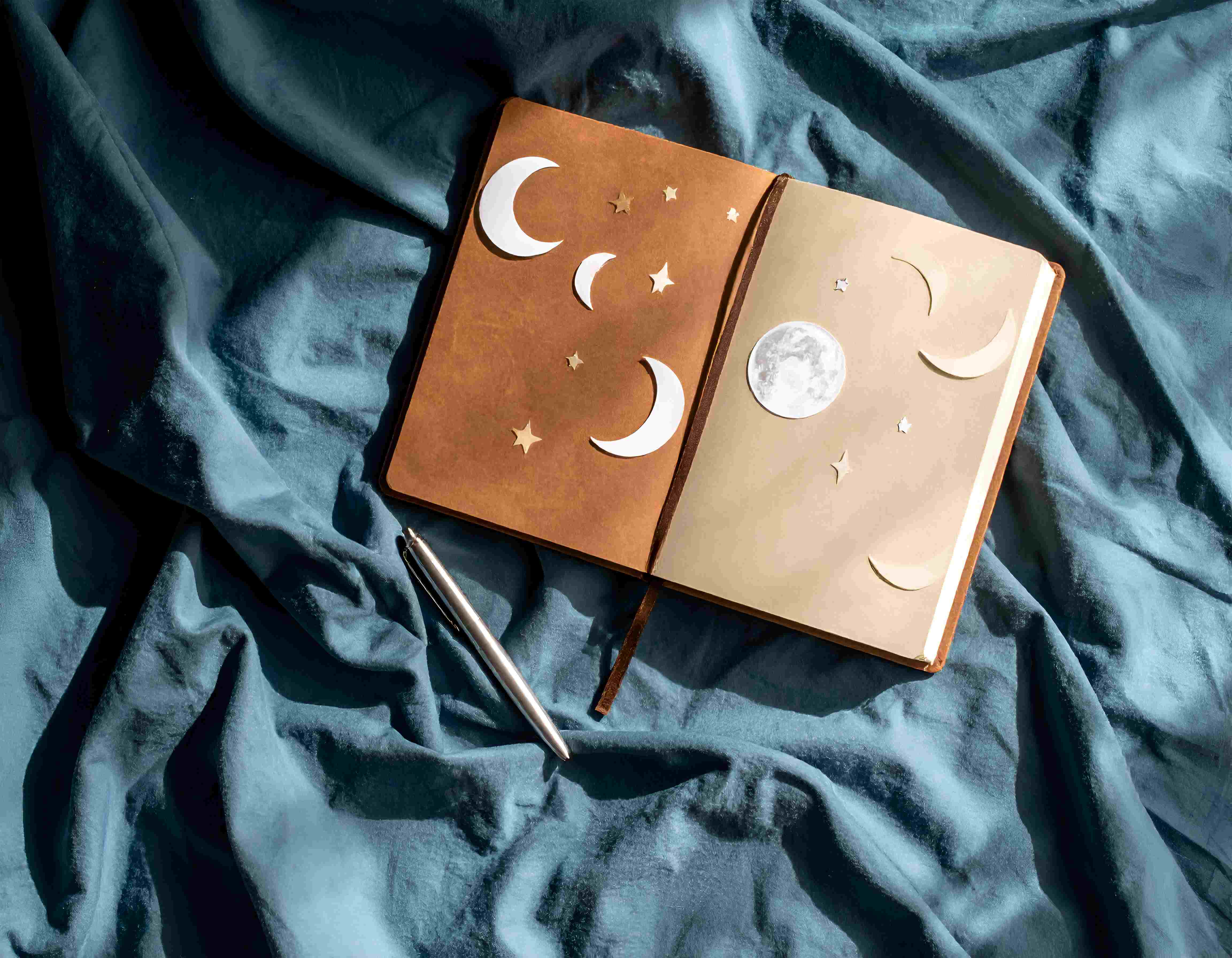 Ein geöffnetes Notizbuch mit Mond- und Sternmotiven liegt auf blauer Bettwäsche neben einem silbernen Stift.