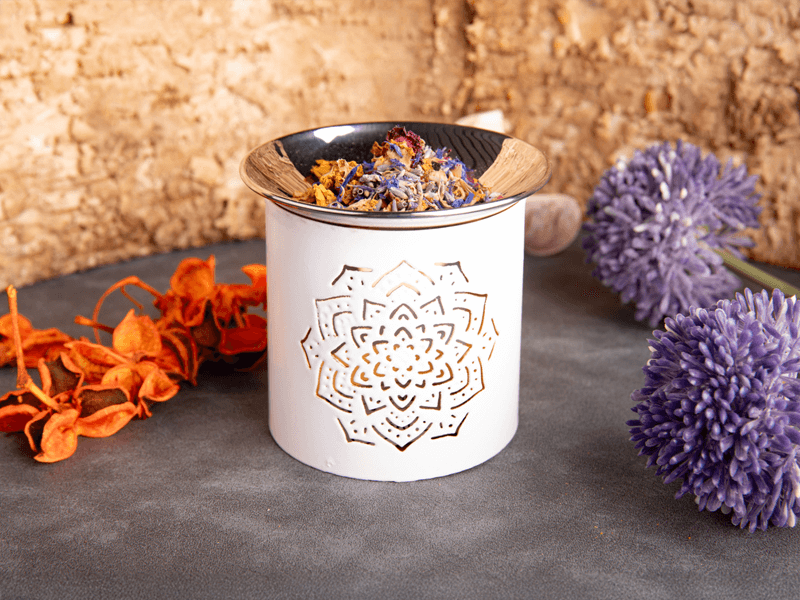 Weißes Räucherstövchen mit filigran ausgestanztem Mandala-Muster, befüllt mit einer Mischung aus getrockneten Blüten in einer Metallschale. 