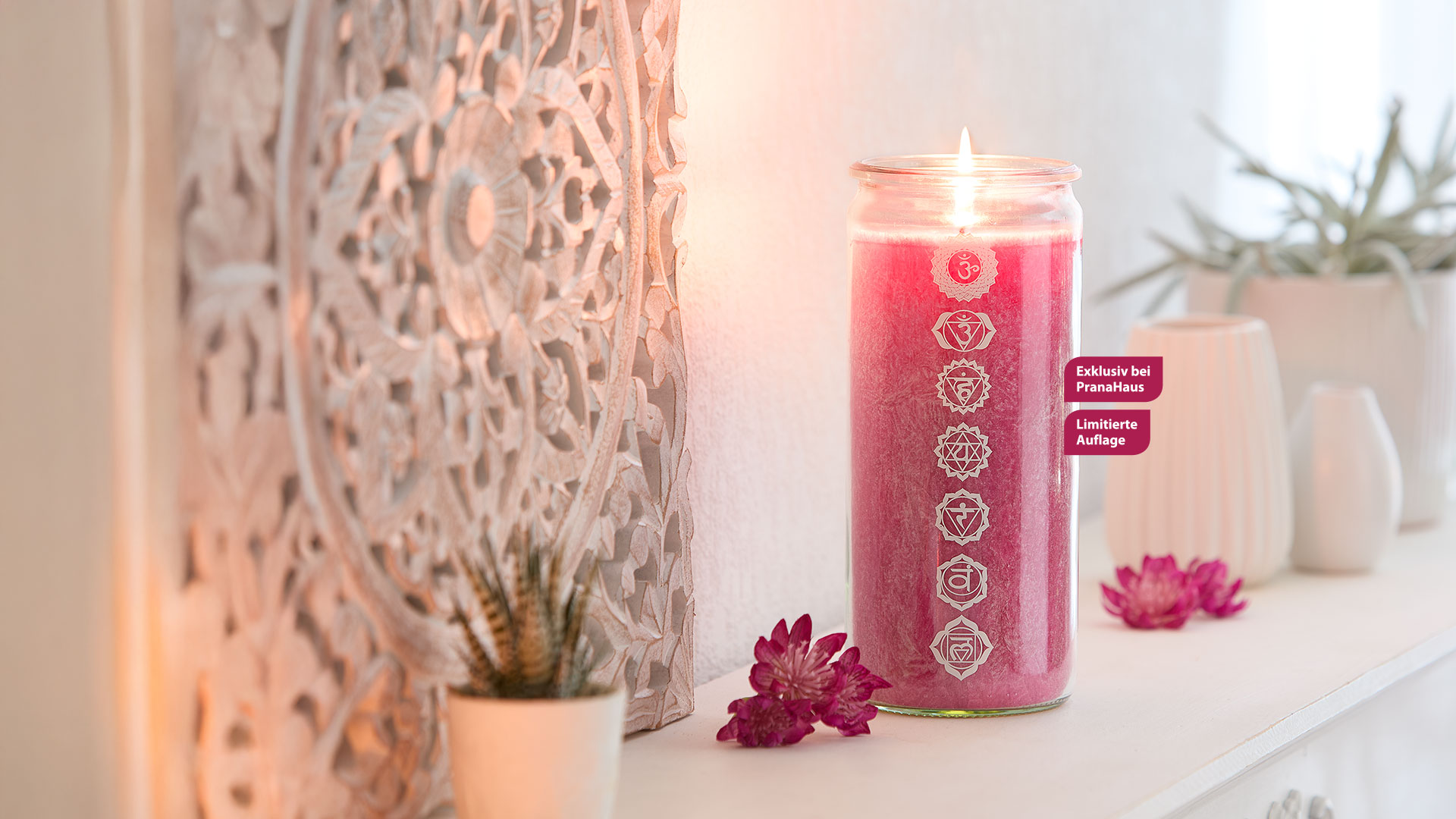 Eine brennende magentafarbene Chakra-Kerze im Glas, bedruckt mit sieben Chakrasymbolen. Im Bild steht: 'Exklusiv bei PranaHaus – Limitierte Auflage'. Die Szene wirkt ruhig und spirituell.
