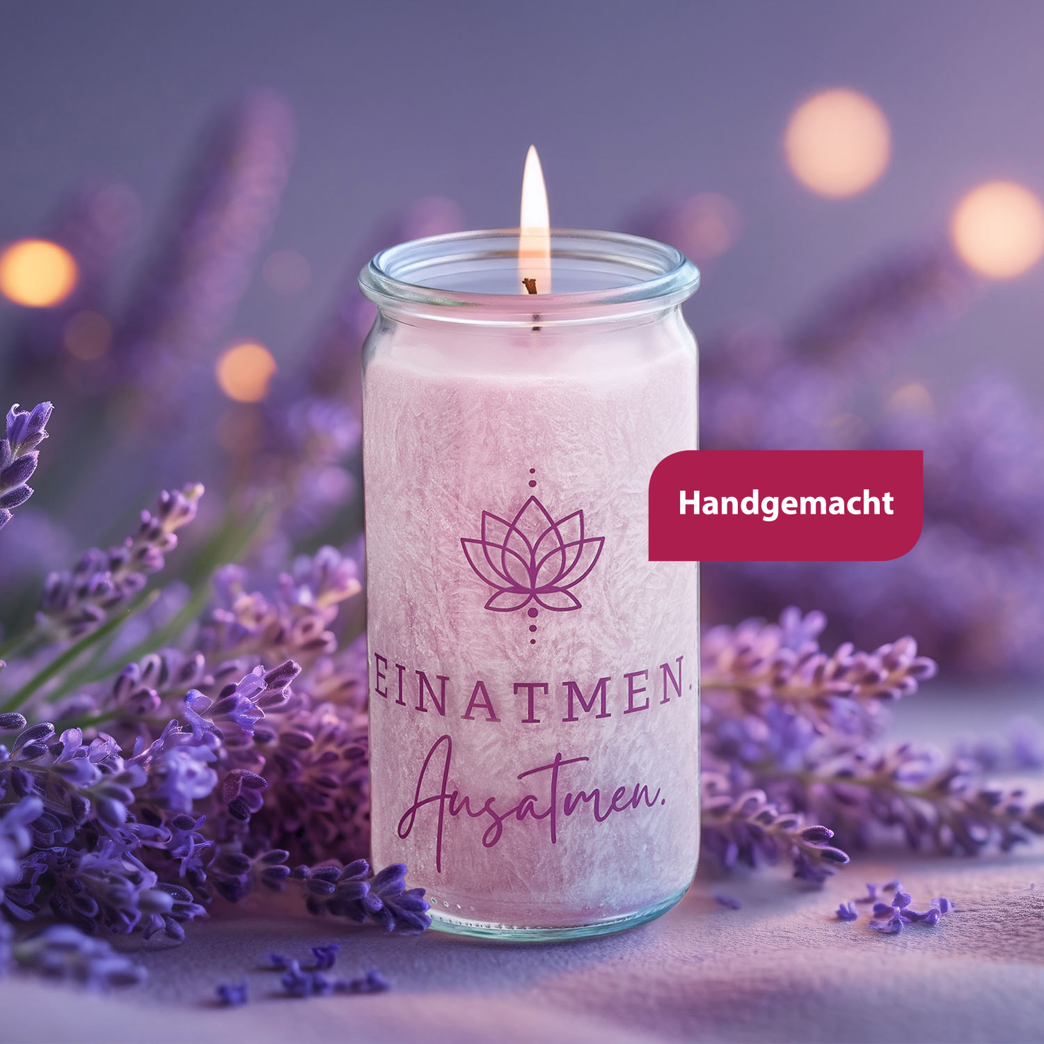 Eine brennende Duftkerze im Glas mit der Aufschrift „Einatmen. Ausatmen.“ steht zwischen lilafarbenen Lavendelblüten vor unscharfem, stimmungsvollem Hintergrund.