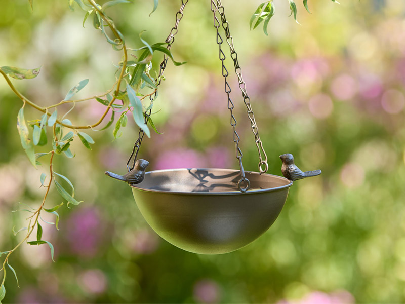 Hängende Metallschale als Vogeltränke mit kleinen Vogelfiguren am Rand, befestigt an Ketten unter einem Ast. Umgeben von zarten Blättern und Blüten entsteht eine ruhige, naturnahe Gartenstimmung.