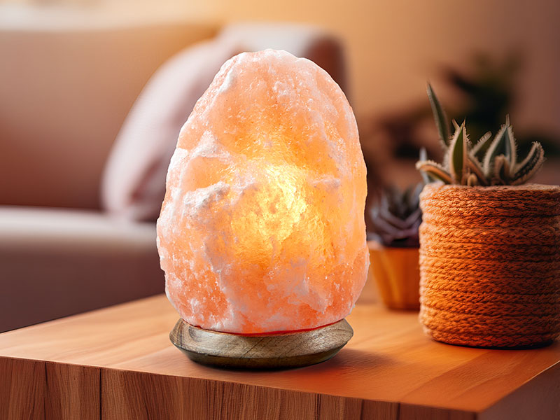 Naturbelassene Salzkristalllampe, die warmes Licht verströmt. Die Lampe steht auf einem Holzsockel auf einem Holztisch in einem gemütlichen Wohnzimmer.