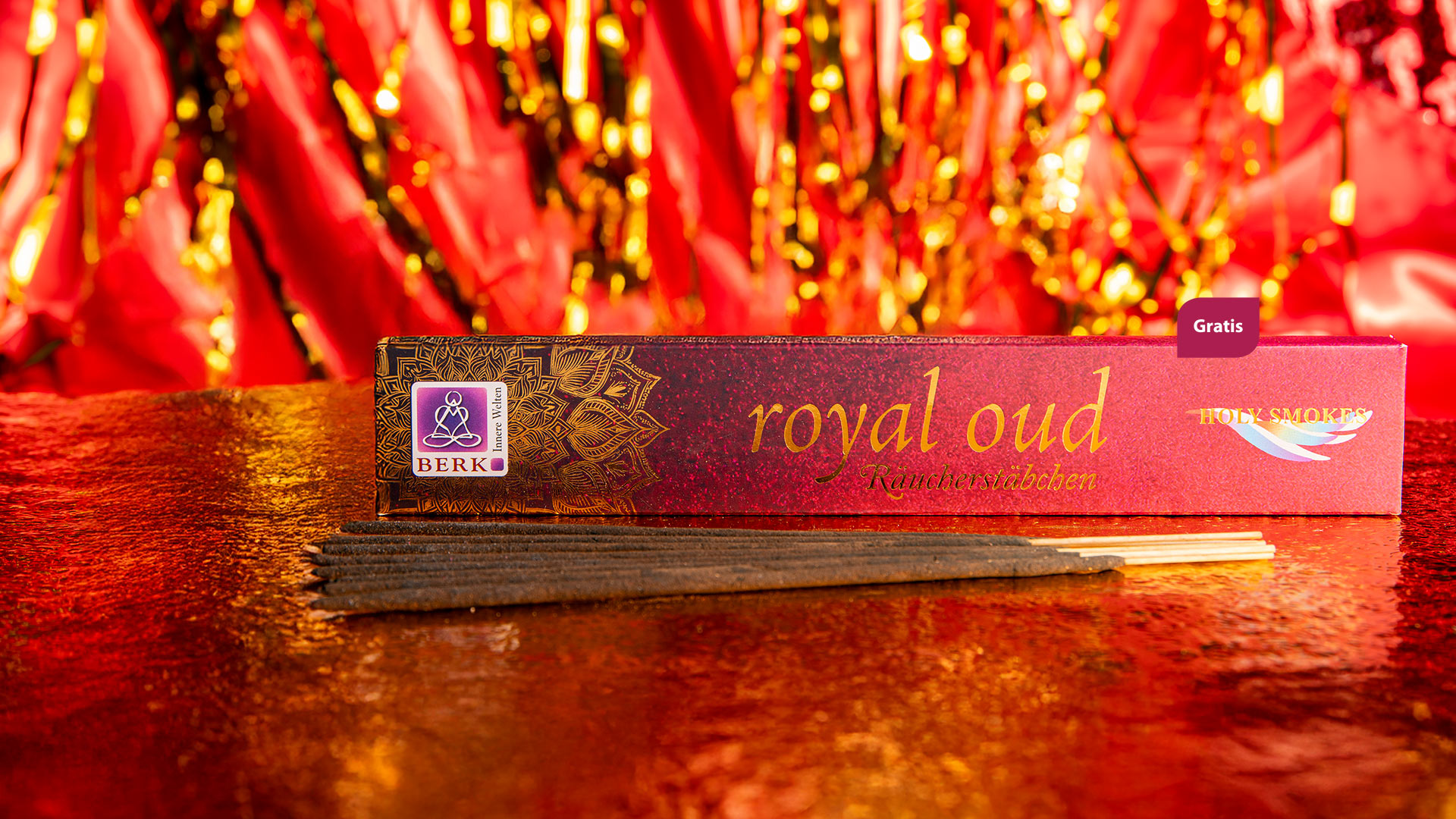 Eine edle Packung Räucherstäbchen der Sorte „Royal Oud“ liegt auf einer glitzernden rot-goldenen Oberfläche. Vor der Verpackung sind mehrere dunkle Räucherstäbchen ausgebreitet.
