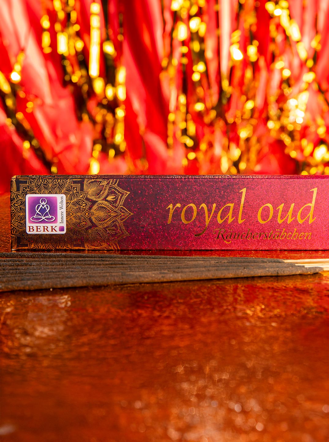 Eine edle Packung Räucherstäbchen der Sorte „Royal Oud“ liegt auf einer glitzernden rot-goldenen Oberfläche. Vor der Verpackung sind mehrere dunkle Räucherstäbchen ausgebreitet.