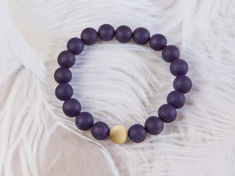 Ein Armband aus matten, dunkelvioletten Perlen liegt auf einem flauschigen Untergrund aus weißen Federn. In der Mitte des Schmuckstücks hebt sich eine helle Weihrauch-Kugel hervor.